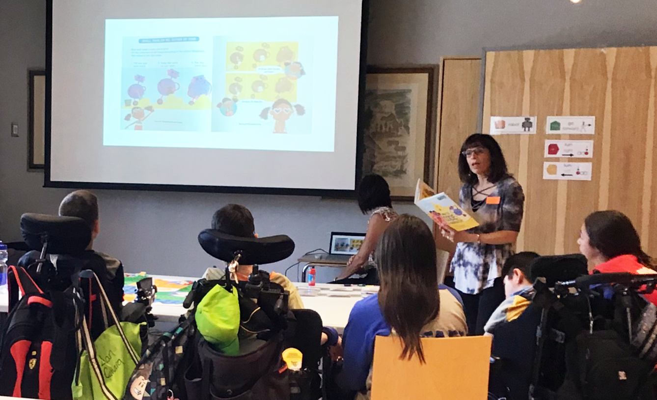 An instructor is reading from a picture book that is also projected on the screen for a group of students sitting on wheelchairs.